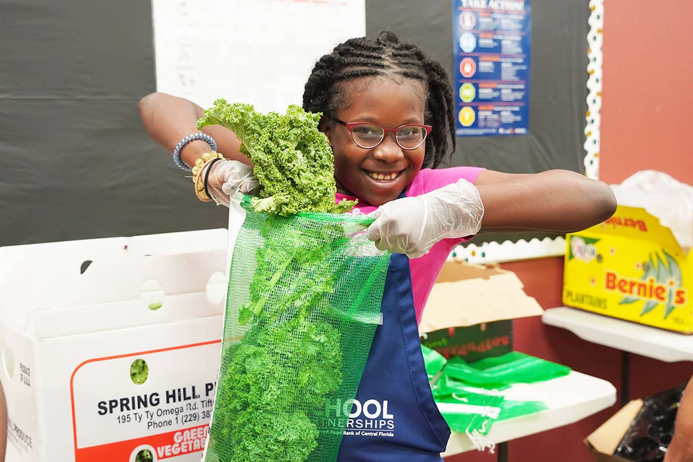A smiling young girl wearing red glasses, gloves, and a blue apron labeled “School Partnerships” holds a bunch of kale while filling a green mesh produce bag. She stands in a classroom setting with produce boxes around her.