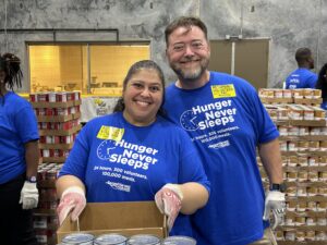 Image of two people smiling while volunteering for Hunger Never Sleeps 24-hour volunteerism event. They are wearing blue t-shirts with the event title "Hunger Never Sleeps" on them and packing food items into boxes.